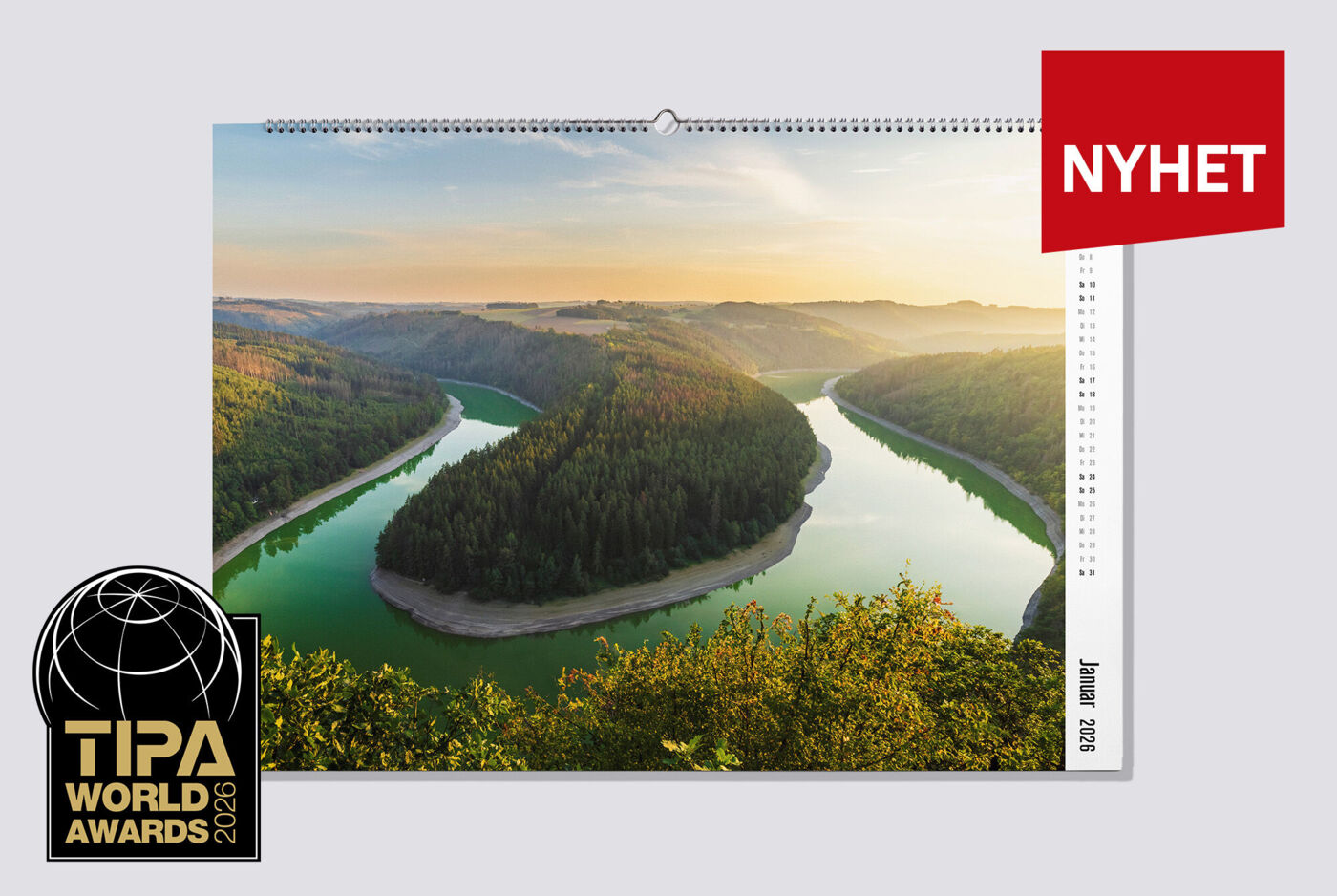 En stor veggkalender i liggende format med et naturbilde av en elv og et skogsområde henger på veggen i en stue. Måned, årstall og ukedager står på høyre side