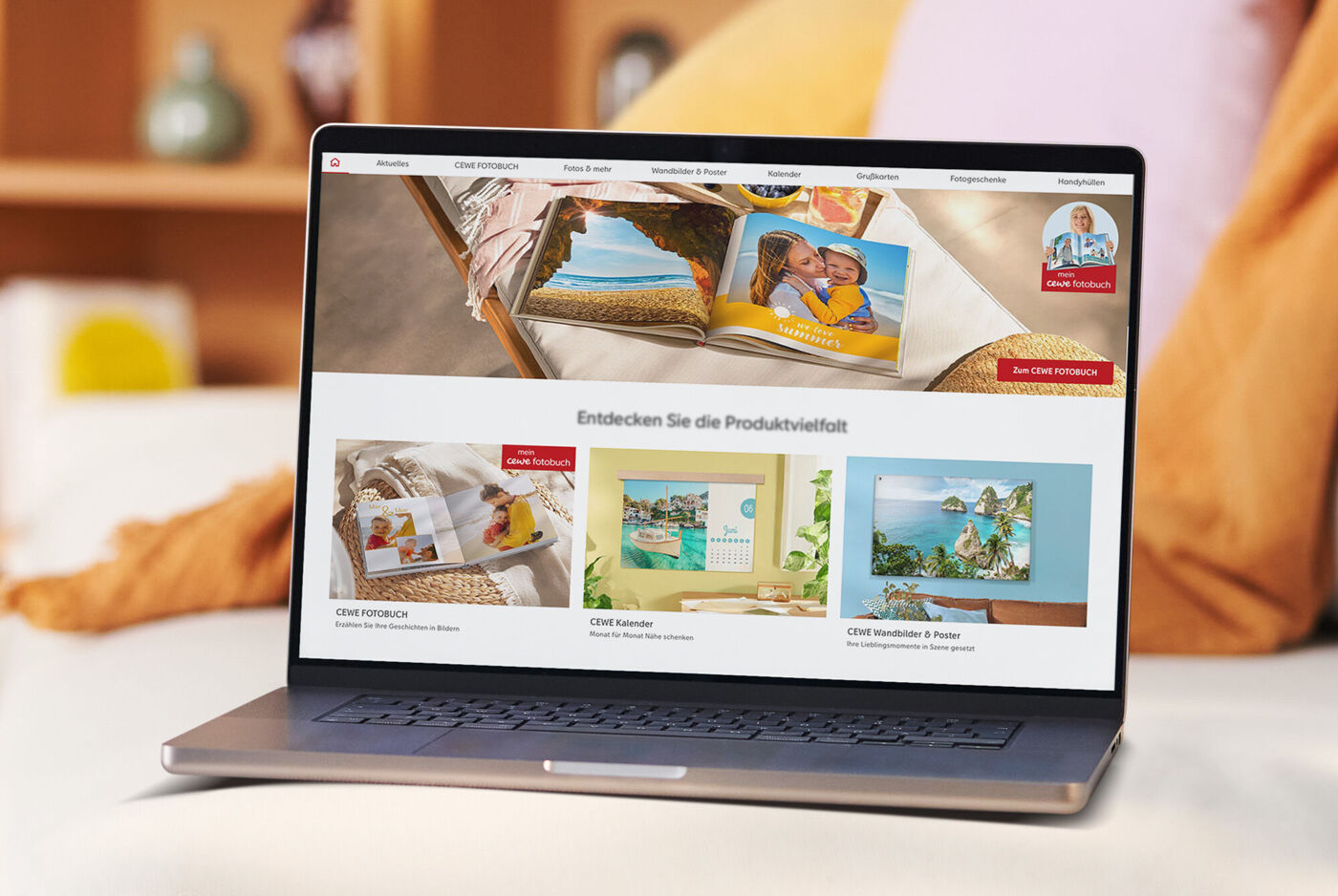 En laptop på en sofa viser CEWE produktspekter med bilder av fotobøker, veggbilder og gratulasjonskort.